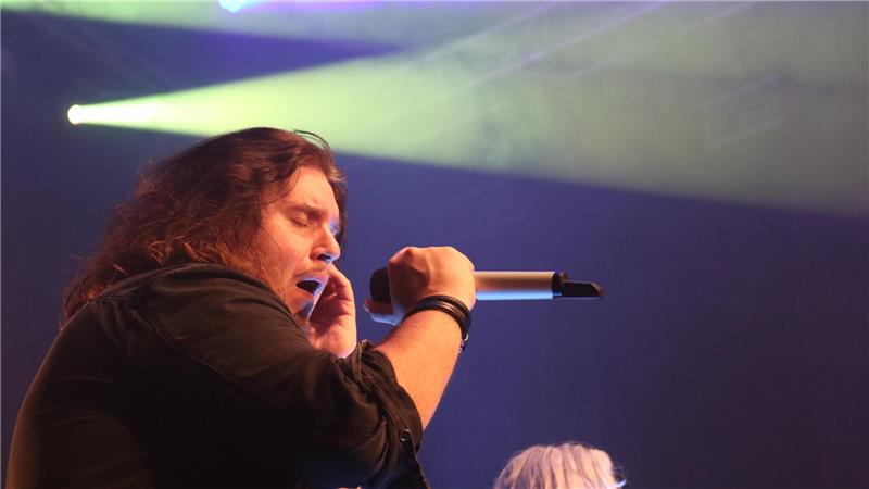 Sänger mit langem Haar hält Mikrofon in der Hand, Bühnenlichtstrahlen im Hintergrund.