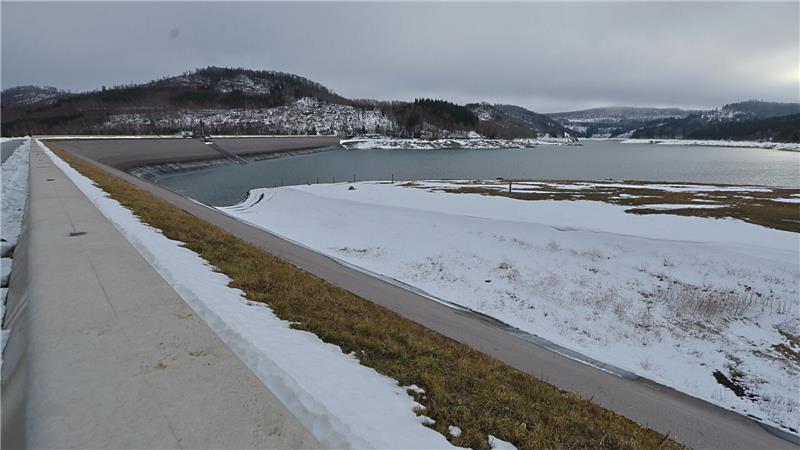 Winterliche Landschaft mit schneebedecktem Damm und See im Hintergrund unter bewölktem Himmel.