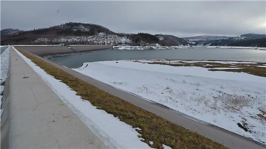 Winterliche Landschaft mit schneebedecktem Damm und See im Hintergrund unter bewölktem Himmel.
