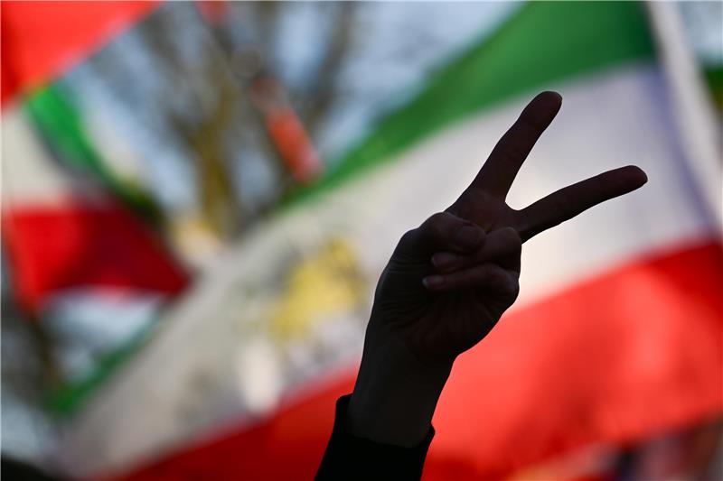 Die Teilnehmerin einer Demo zeigt vor einer iranischen Flagge das Victory-Zeichen mit ihrer Hand.