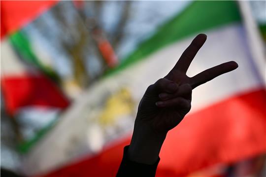 Die Teilnehmerin einer Demo zeigt vor einer iranischen Flagge das Victory-Zeichen mit ihrer Hand.
