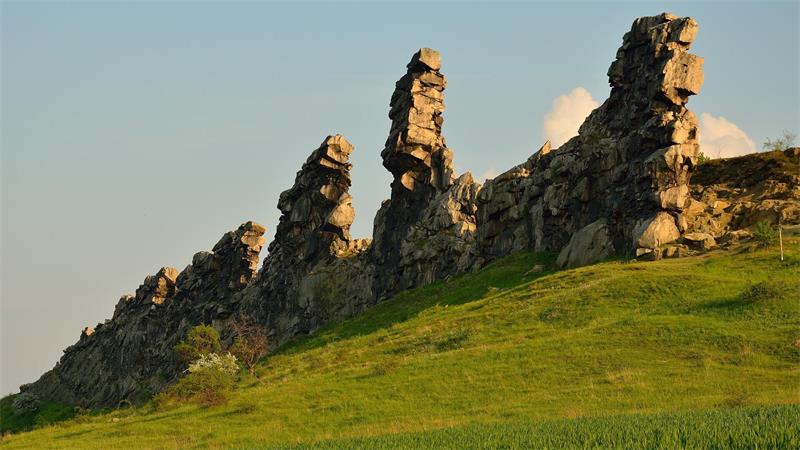 Felsformation mit mehreren hohen, schmalen, unregelmäßig geformten Steinsäulen auf einem grasbewachsenen Hügel unter klarem Himmel.