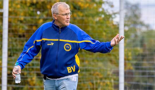 Mann in blau-gelber Sportjacke und Jeans hält Wasserflasche und gestikuliert auf einem Fußballfeld
