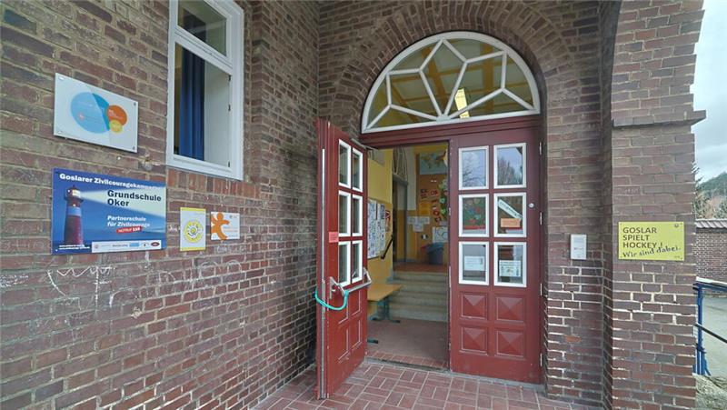 Offene rote Doppeltür mit Glasfenstern in einem Backsteingebäude, links Schilder an der Wand, im Inneren Treppe und gelbe Sitzbank sichtbar.