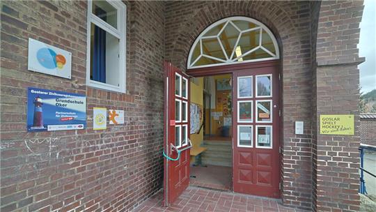 Offene rote Doppeltür mit Glasfenstern in einem Backsteingebäude, links Schilder an der Wand, im Inneren Treppe und gelbe Sitzbank sichtbar.