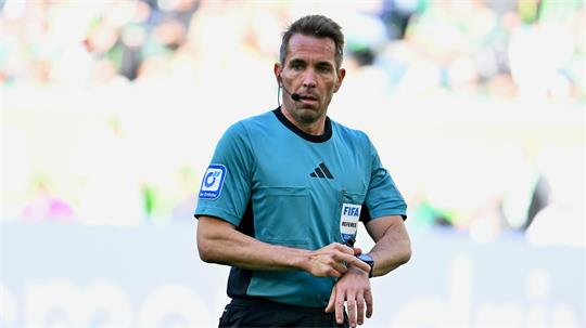 Schiedsrichter in blauem Trikot mit FIFA-Logo und Mikrofon am Ohr blickt auf die Uhr an seinem Handgelenk