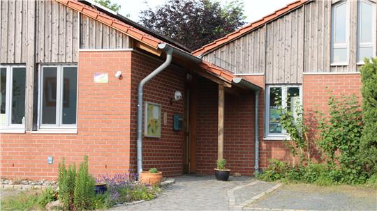 Eingangsbereich eines eingeschossigen Hauses mit rotem Ziegelfundament, Holzverkleidung und einem überdachten Hauseingang mit zwei Pflanzen in Töpfen