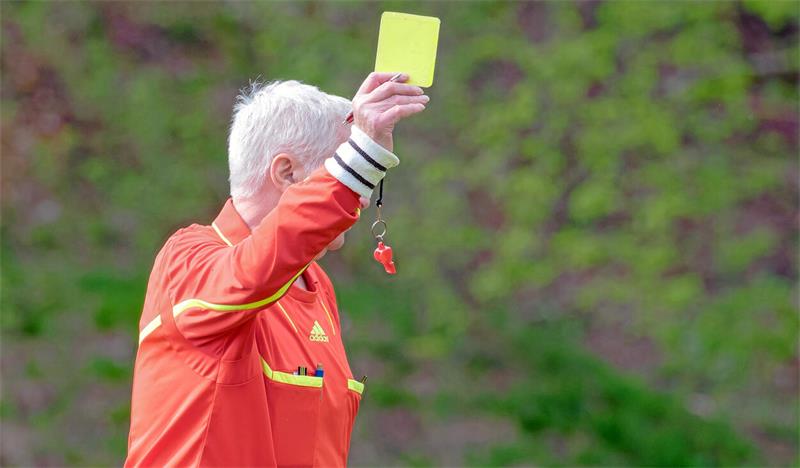 Schiedsrichter mit grauen Haaren in orangefarbener Jacke hält Gelbe Karte hoch und trägt Pfeife um den Arm.