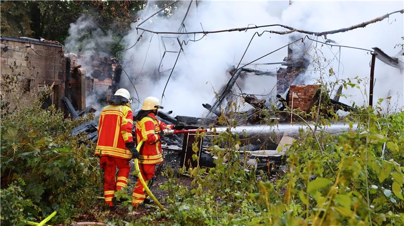 Zwei Feuerwehrleute in Schutzkleidung löschen ein brennendes Gebäude, umgeben von Rauch und Vegetation