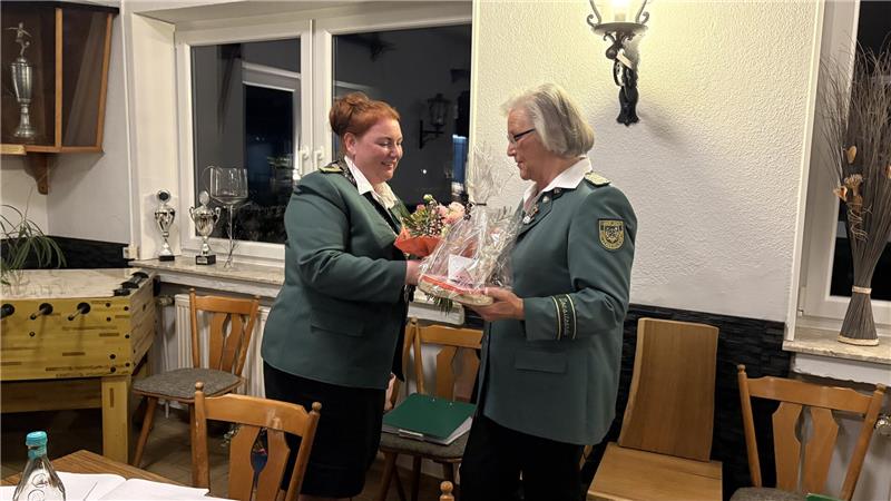 Zwei Frauen in grünen Uniformjacken übergeben sich in einem Raum mit Holzmöbeln und Pokalen ein Geschenkpaket.
