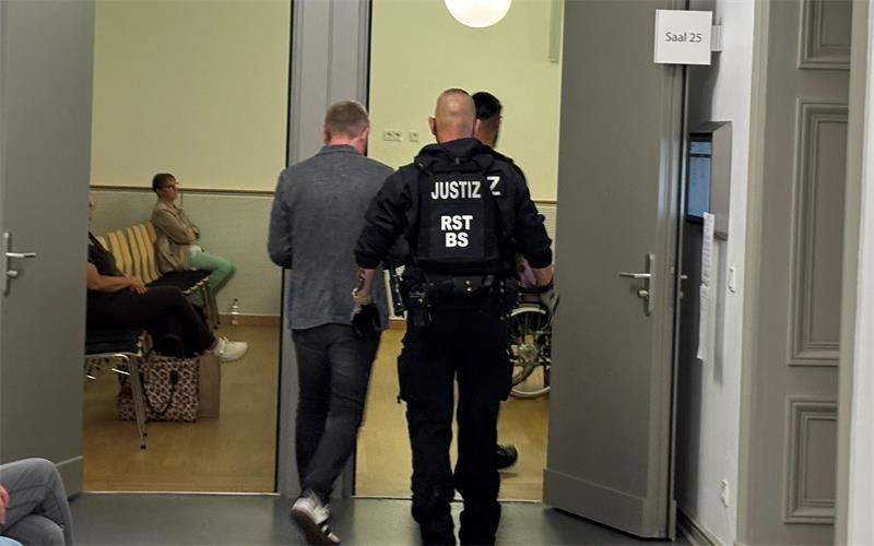 Ein Justizbeamter führt einen Mann durch eine geöffnete Tür in einen Raum mit Sitzgelegenheiten, in dem eine weitere Person sitzt.