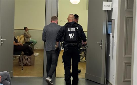 Ein Justizbeamter führt einen Mann durch eine geöffnete Tür in einen Raum mit Sitzgelegenheiten, in dem eine weitere Person sitzt.