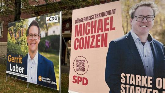 Zwei nebeneinander stehende Wahlplakate mit Porträtfotos von Männern in Anzügen, links mit dem Namen Gerrit Lober und CDU-Logo, rechts mit dem Namen Michael Conzen, SPD-Logo und dem Text 'Bürgermeisterkandidat'.