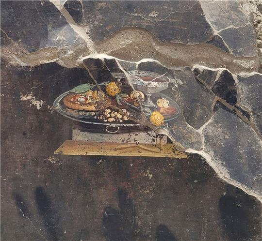 Die Wand eines pompejanischen Hauses mit einem Fresko, das einen Tisch mit Speisen darstellt, darunter auch eine Art Pizza.
