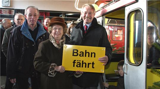Gruppe von Menschen steigt in eine Seilbahn ein, zwei Personen halten ein gelbes Schild mit der Aufschrift 'Bahn fährt'
