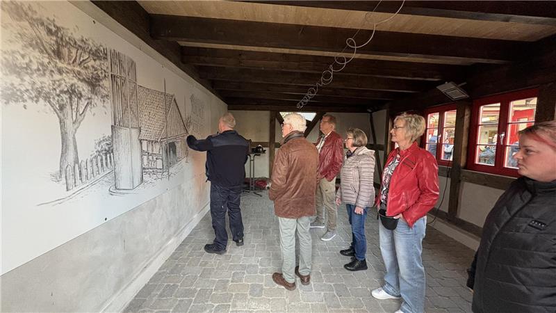Pferdespritze nimmt ihren Platz im Kleinen Spritzenhaus ein Fünf Personen stehen in einem Raum mit Holzbalkendecke vor einer großen schwarz-weißen Wandzeichnung, eine Person zeigt darauf