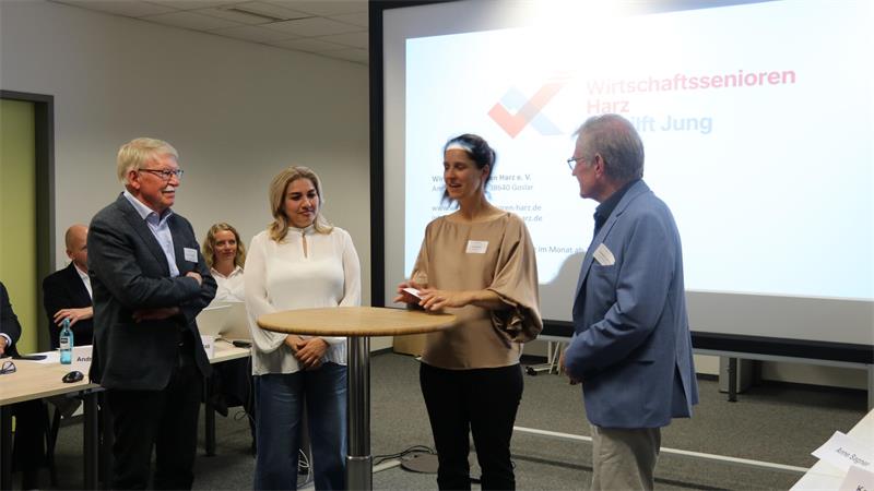 Vier Personen stehen um einen Stehtisch in einem Konferenzraum, im Hintergrund eine Projektionsleinwand mit unscharf lesbarem Text.