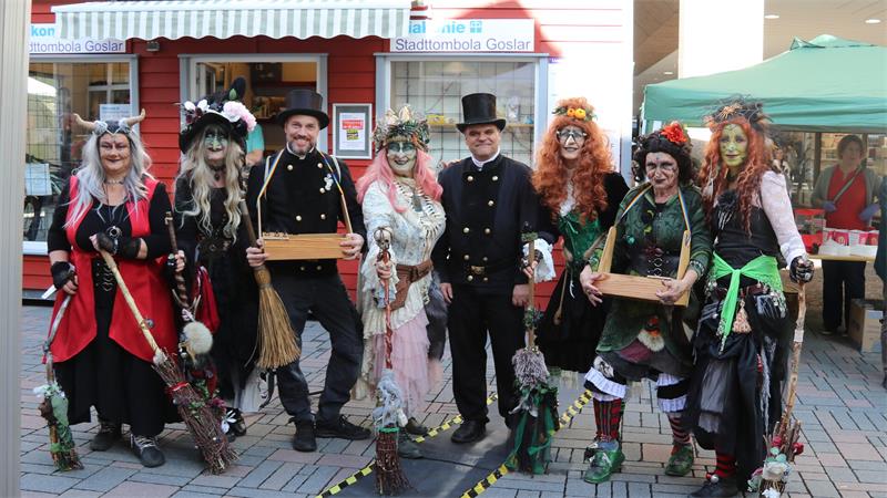 Gruppe von Menschen in fantasievollen Hexenkostümen mit Besen vor einem Stand mit dem Schild 'Stadttombola Goslar' auf einem gepflasterten Platz