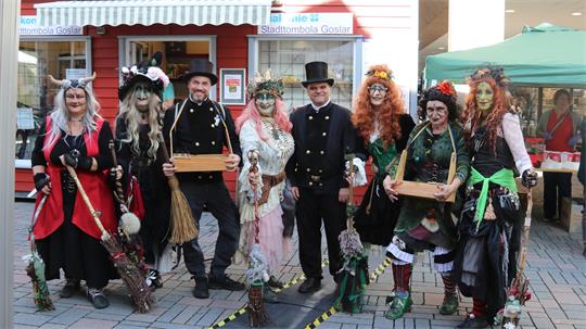Gruppe von Menschen in fantasievollen Hexenkostümen mit Besen vor einem Stand mit dem Schild 'Stadttombola Goslar' auf einem gepflasterten Platz