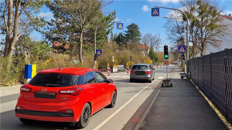 Rote und graue Autos fahren auf einer Straße mit Fußgängerüberweg und grünem Ampelsignal bei klarem Himmel.