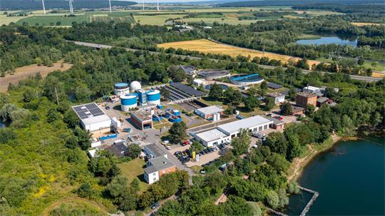 Luftaufnahme einer Industrieanlage mit mehreren Gebäuden, Tanks und Solarpaneelen, umgeben von Wald, Feldern und einem See