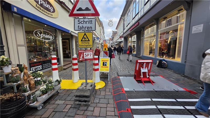 Fußgängerzone mit Kopfsteinpflaster, mehreren Verkehrsschildern und Absperrungen, Geschäfte links und rechts, Menschen im Hintergrund.