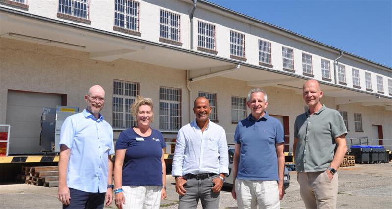 Fünf Personen stehen vor einem hellen Gebäude mit mehreren Fenstern und Müllcontainern und blicken in die Kamera