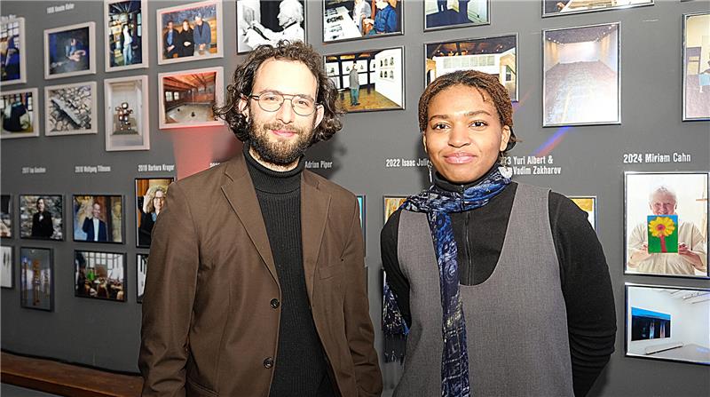 Ein Mann und eine dunkelhäutige Frau blicken vor einer Bildergalerie in die Kamera.