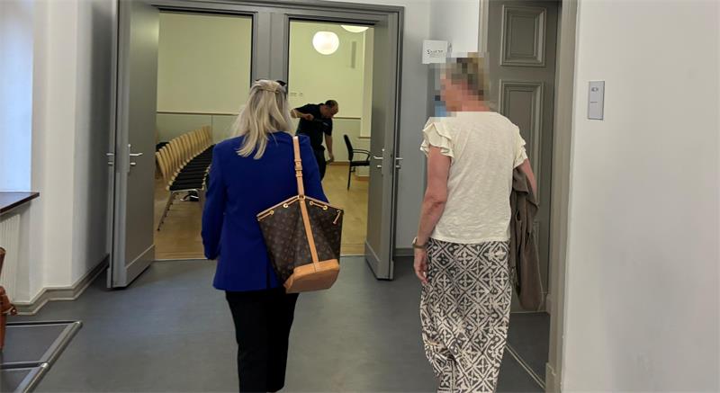 Zwei Frauen gehen einen Flur entlang in Richtung eines Raums mit Stuhlreihen, eine trägt eine blaue Jacke und eine Tasche, die andere eine weiße Bluse und gemusterte Hose