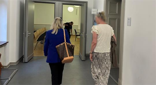 Zwei Frauen gehen einen Flur entlang in Richtung eines Raums mit Stuhlreihen, eine trägt eine blaue Jacke und eine Tasche, die andere eine weiße Bluse und gemusterte Hose