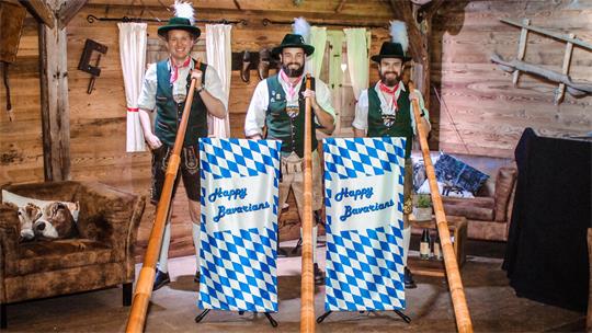 Drei Männer in traditioneller bayerischer Tracht stehen in einer Holzhütte und halten lange Alphörner vor zwei blau-weißen Fahnen mit der Aufschrift 'Happy Bavarian'