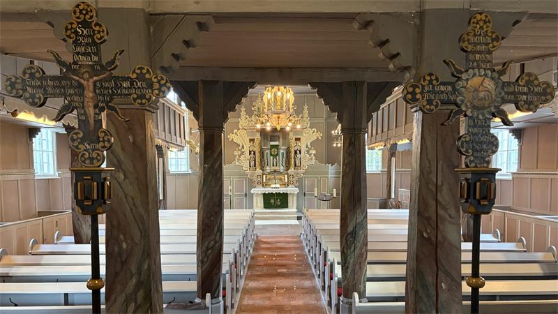 Innenraum einer Kirche mit hölzernen Bänken, verzierten Holzkreuzen im Vordergrund und einem goldverzierten Altar im Hintergrund.