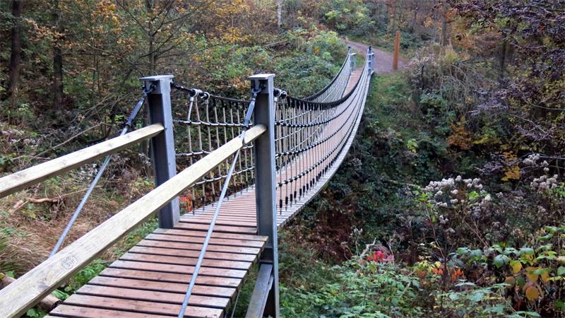 Hängebrücke aus Holz und Metall mit Seilgeländer führt durch bewaldetes Tal.