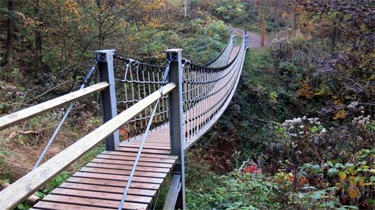 Hängebrücke aus Holz und Metall mit Seilgeländer führt durch bewaldetes Tal.