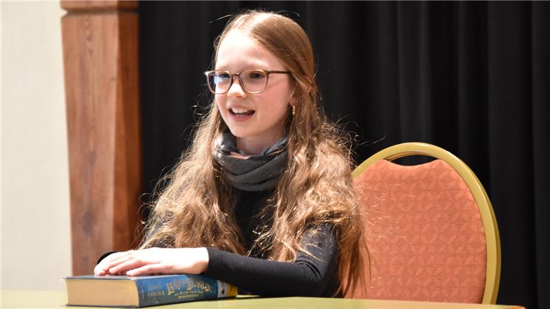 Mädchen mit langen, welligen Haaren sitzt an einem Tisch mit einem Buch. Im Hintergrund ein orangefarbener Stuhl.