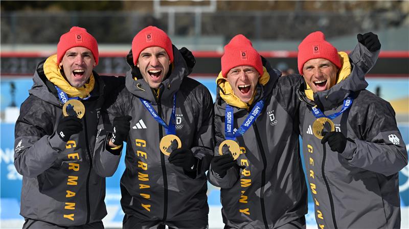Vier Athleten in schwarzen Jacken mit gelber Aufschrift "GERMANY" und roten Mützen posieren mit Goldmedaillen vor einer olympischen Kulisse