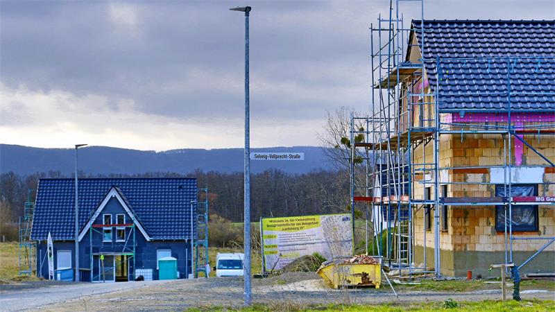 Zwei im Bau befindliche Häuser an einer Straße mit Baustellengerüst und Baustellenschildern unter bewölktem Himmel