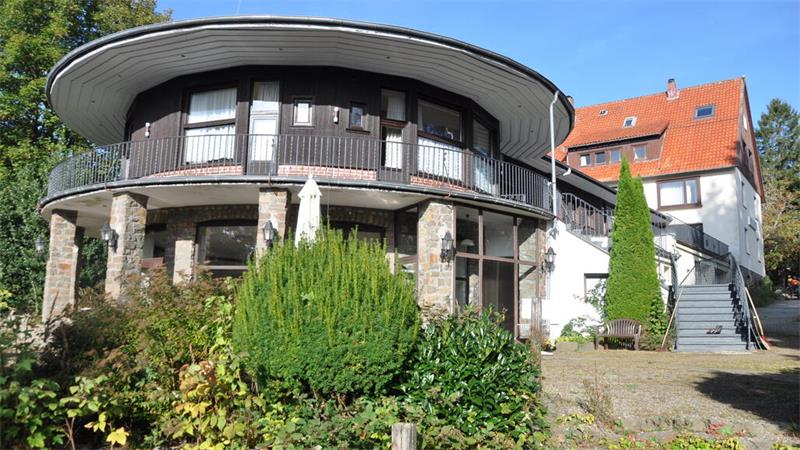 Rundes zweistöckiges Haus mit Balkon, Säulen und umlaufendem Dach, umgeben von Büschen und Bäumen