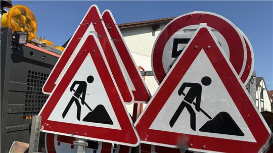 Mehrere dreieckige Baustellenschilder mit rotem Rand und schwarzer Piktogrammfigur, die mit Schaufel arbeitet, vor einem Fahrzeug und Häusern im Hintergrund