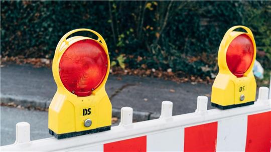 Gelbe Warnleuchten mit roten Reflektoren auf einer rot-weißen Absperrung vor einem Gehweg und Hecke im Hintergrund.