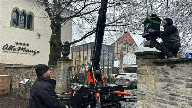 Zwei Personen bedienen eine kleine Raupenkranmaschine, um eine Statue auf einem steinernen Mauersockel vor einem Gebäude mit der Aufschrift 'Alte Münze' zu platzieren.