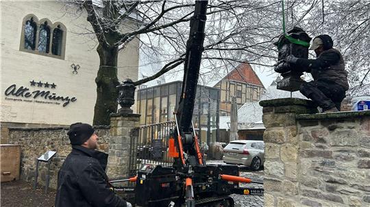 Zwei Personen bedienen eine kleine Raupenkranmaschine, um eine Statue auf einem steinernen Mauersockel vor einem Gebäude mit der Aufschrift 'Alte Münze' zu platzieren.