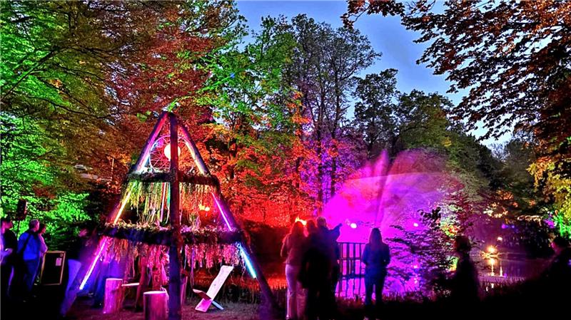 Menschen stehen abends im Wald vor einer mit bunten Lichtern beleuchteten Holzkonstruktion und einem beleuchteten Wasserbrunnen