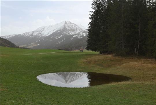 Die im Dunst liegenden Alpen spiegeln sich in einem Weiher. Der Saharastaub-Höhepunkt ist überschritten.