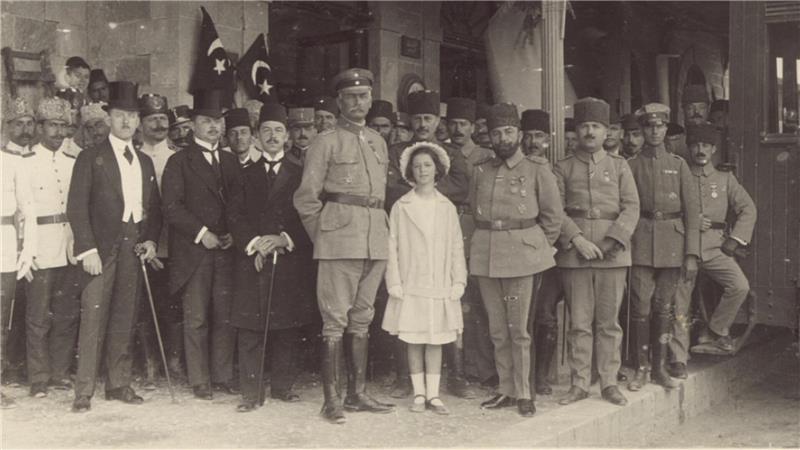 Gruppenaufnahme von Männern in Militär- und Ziviluniformen sowie ein Mädchen in hellem Kleid vor einem Gebäude mit Säulen