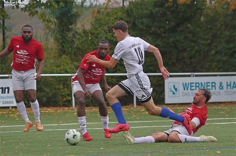 Fußballspieler in Aktion auf einem Spielfeld mit grünem Kunstrasen, ein Spieler in weißem Trikot läuft am Ball vorbei, während ein Spieler in rotem Trikot auf dem Boden liegt und andere Spieler in roten Trikots aufmerksam zuschauen.