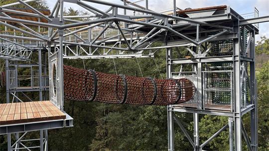 Metallgerüst mit hängender, röhrenförmiger Kletterbrücke aus Netzmaterial in einem Waldgebiet.