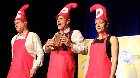 Zwei Männer und eine Frau stehen auf einer Bühne nebeneinander in roten Schürzen und roten Zwergenzipfelmützen mit den Buchstaben: SPD.
