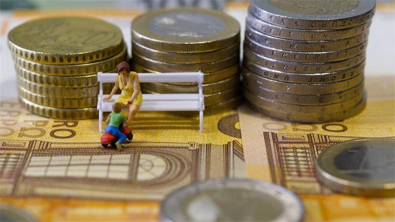 Miniaturfiguren einer sitzenden Frau und eines knienden Kindes vor gestapelten Münzen auf Geldscheinen