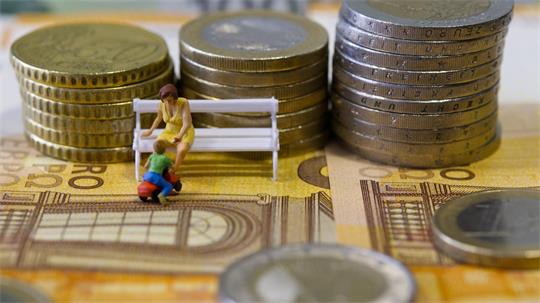 Miniaturfiguren einer sitzenden Frau und eines knienden Kindes vor gestapelten Münzen auf Geldscheinen